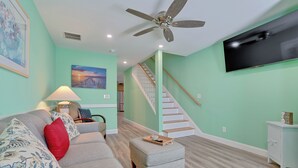 A living room with a grey sofa, red and blue pillows, a grey ottoman, a ceiling fan, a flat-screen TV, a staircase, and a light blue wall. - Across the street from the beach with EV Charger.  Unit A in Duplex (Old Orchard Beach)