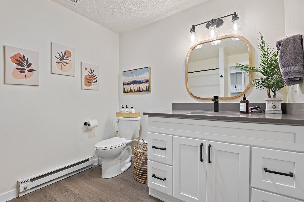 A modern bathroom with a toilet, white cabinets, a mirror with lights, and wall art.