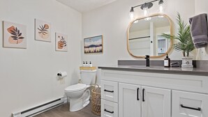 A modern bathroom with a toilet, white cabinets, a mirror with lights, and wall art. - Upscale Rockwood Boho Townhouse (Spokane)