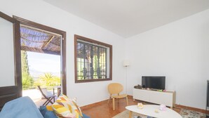 A living room with a blue sofa, a small white table, a TV on a white stand, and a window with a view of greenery. - Cubo's La Maison de Mily / fireplace / private pool / free wifi (Coín)