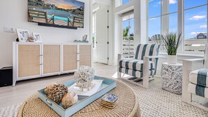 A spacious living room with a large window, a view of a cityscape, a decorative wall piece, and a wicker coffee table. - Beach Craft TC 8202 (Port Aransas)