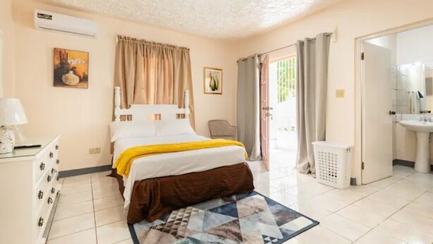 Un dormitorio con cama, cómoda, ventana con cortinas, baño con lavamanos y espejo, y una puerta que da al exterior.