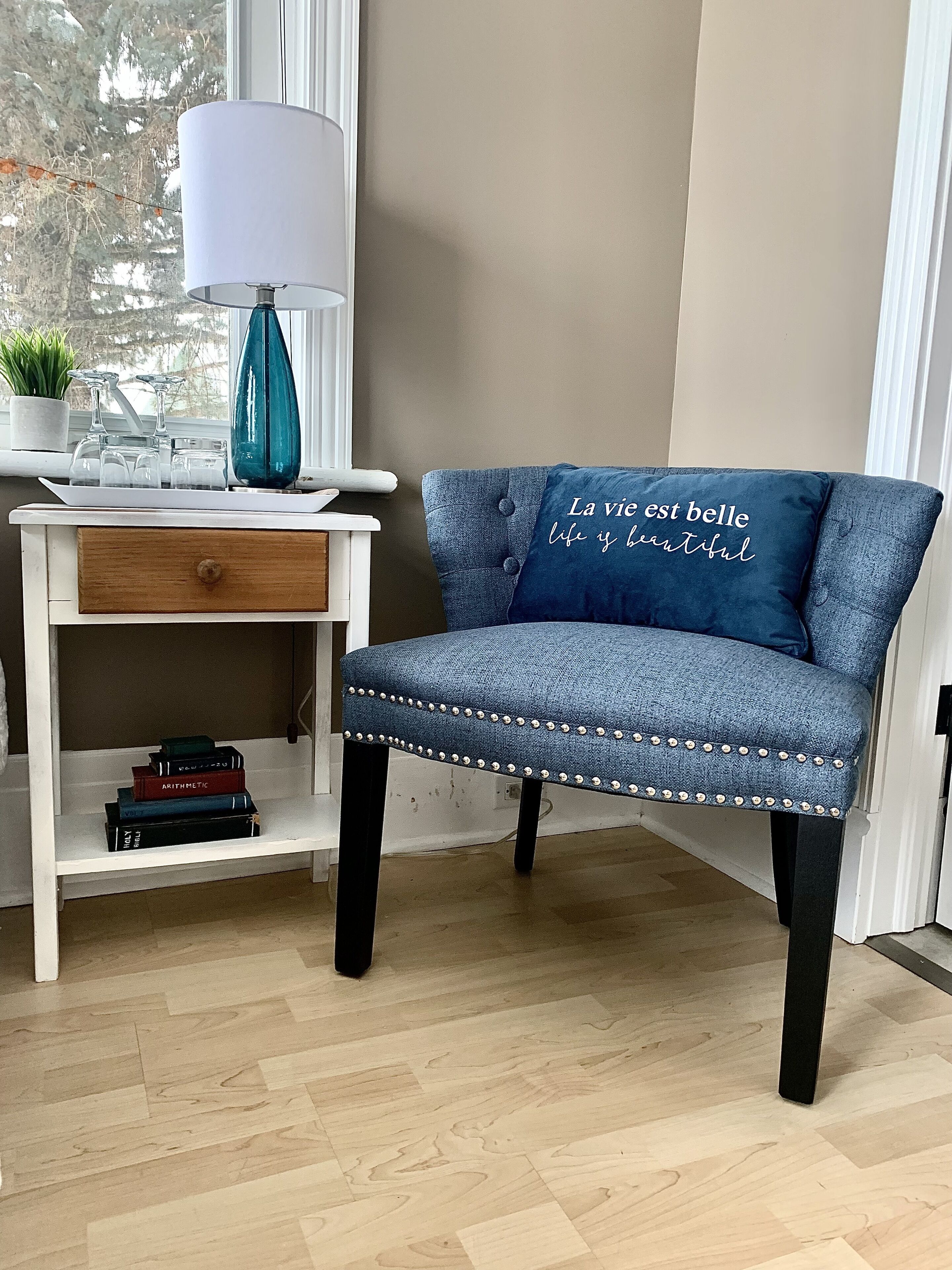 A blue upholstered chair with a cushion that reads "La vie est belle life is beautiful" in white cursive text, positioned next to a white desk with a lamp and a small plant.
