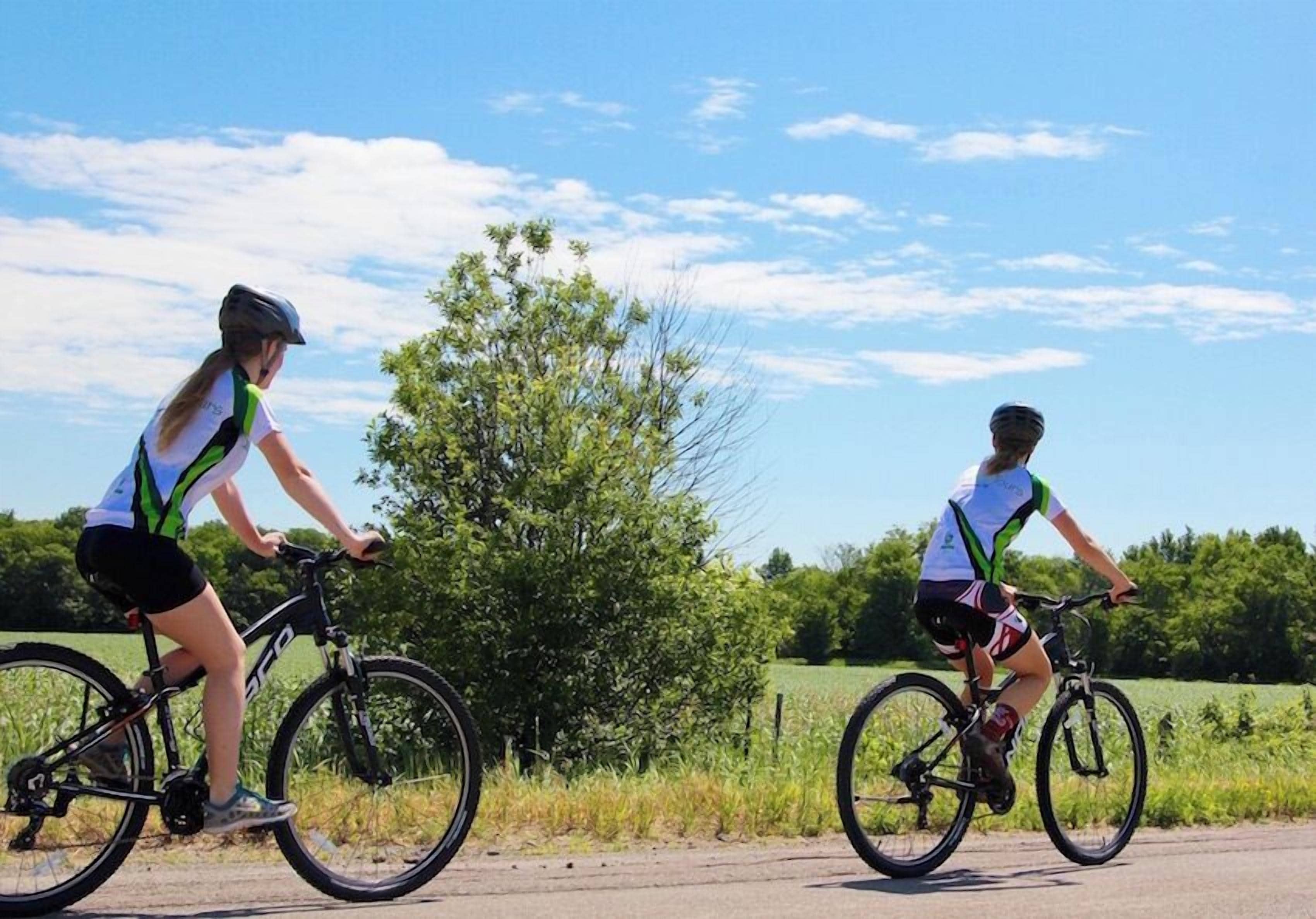 Two cyclists on a paved path with a rural landscape in the background.