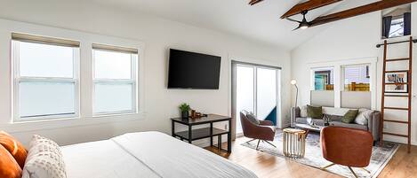 A spacious bedroom with a large bed, a desk, a chair, and a TV. There are windows and a balcony with a view.