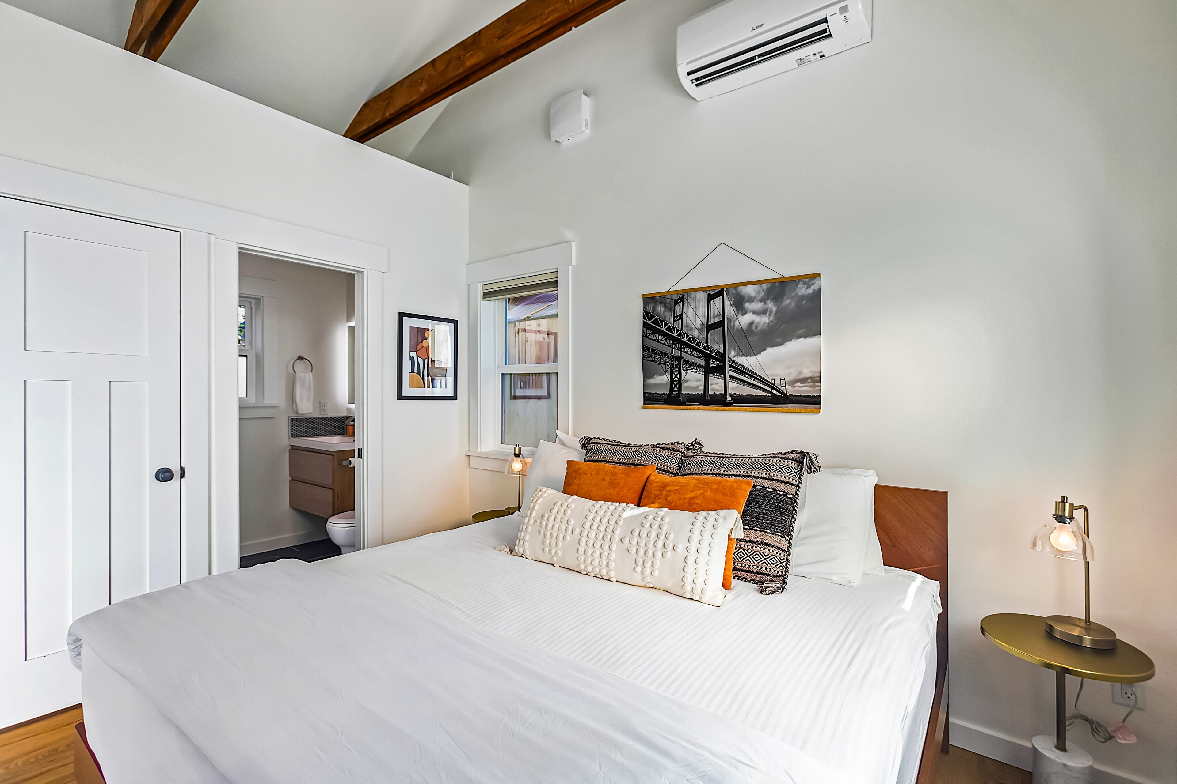A bedroom with a large bed, white bedding, a bedside table with a lamp, a framed picture on the wall, and an air conditioning unit.