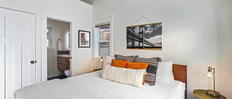 A bedroom with a large bed, white bedding, a bedside table with a lamp, a framed picture on the wall, and an air conditioning unit.