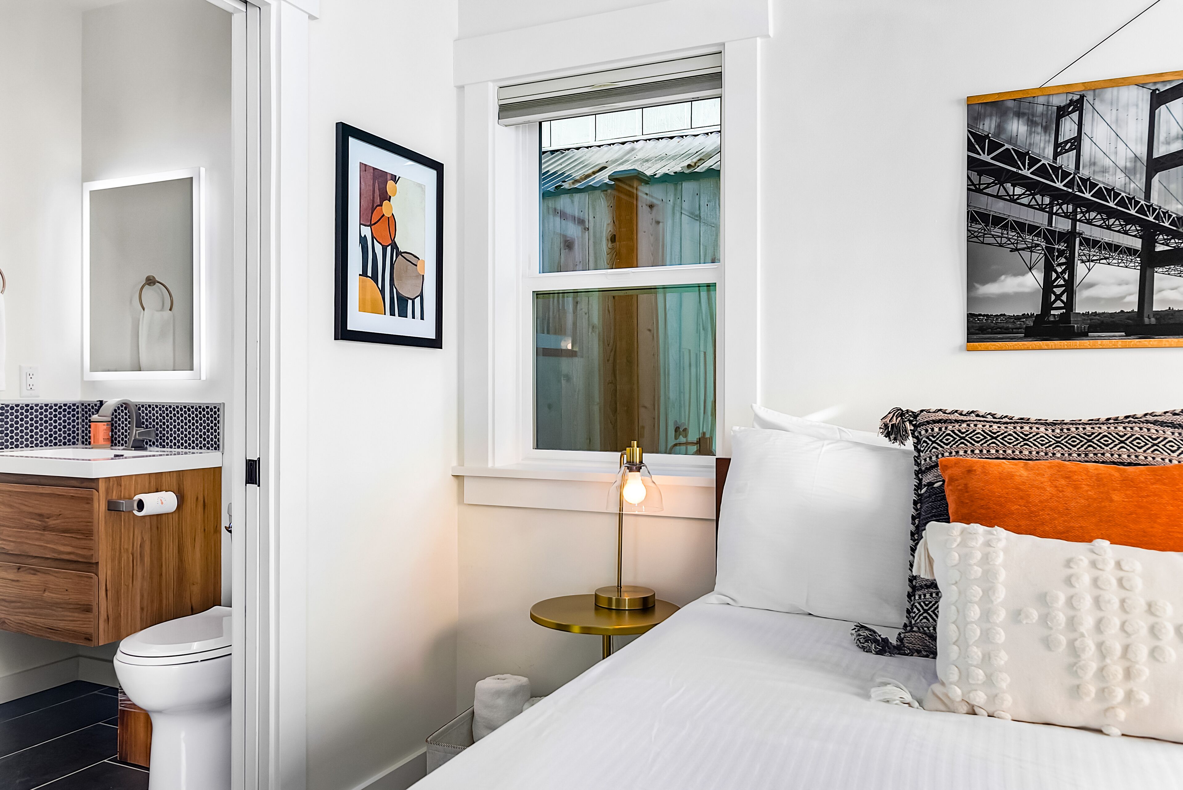 Un dormitorio con cama, una mesita de noche con lámpara, una ventana, un cuadro y un baño con inodoro y lavabo.