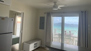 A room with a kitchenette, a ceiling fan, a bench, and a large sliding glass door leading to a balcony with a view of the ocean.