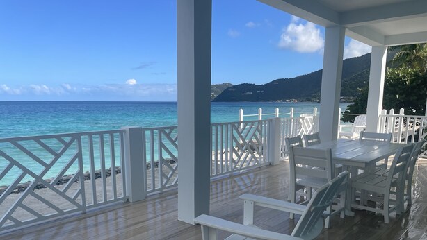 A covered outdoor seating area with white furniture, a wooden deck, and a view of the ocean and hills.