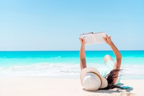 A person lying on a beach with a book, wearing a hat, and a striped shirt. - The Blue Pearl (Miramar Beach)