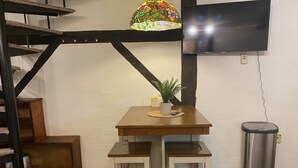 Ein kleiner Essbereich mit einem Holztisch, zwei weißen Stühlen, einer Topfpflanze und einem an der Wand befestigten Fernseher.