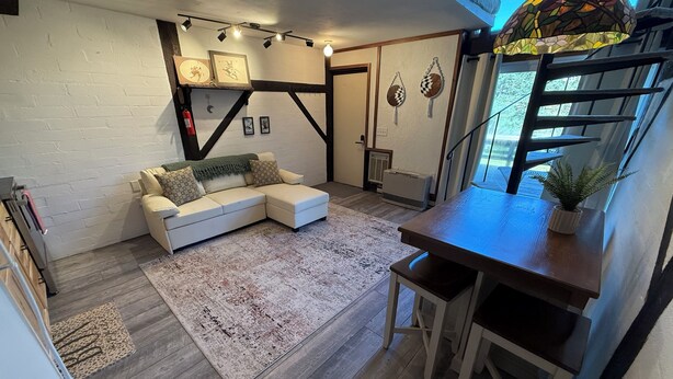Ein gemütliches Wohnzimmer mit einem weißen Modulsystem, einem Holztisch und einem gemusterten Teppich. Eine Treppe führt zu einer oberen Ebene, und ein Fenster bietet Blick auf viel Grün draußen.