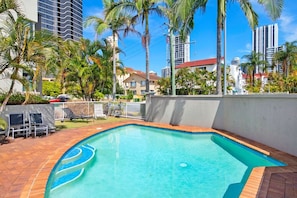 A swimming pool with a curved edge, surrounded by palm trees and a white wall. - Broadbeach Oasis- 2 Bedroom Apartment in Britannia (Broadbeach)