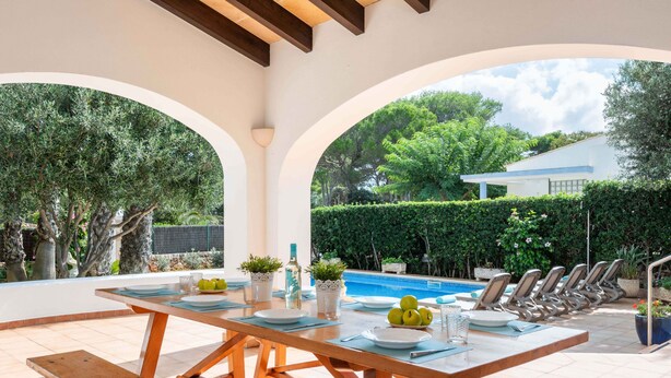 Un espace repas aménagé au bord de la piscine, avec une table en bois pouvant accueillir six personnes, offrant une vue sur la piscine et un jardin.