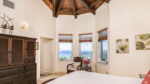 A bedroom with a large bed, wooden dresser, two windows with blinds, a chair, and tropical-themed wall art. - View Come True Villa, Panoramic Views from a Wraparound Balcony in St. John (St. John)