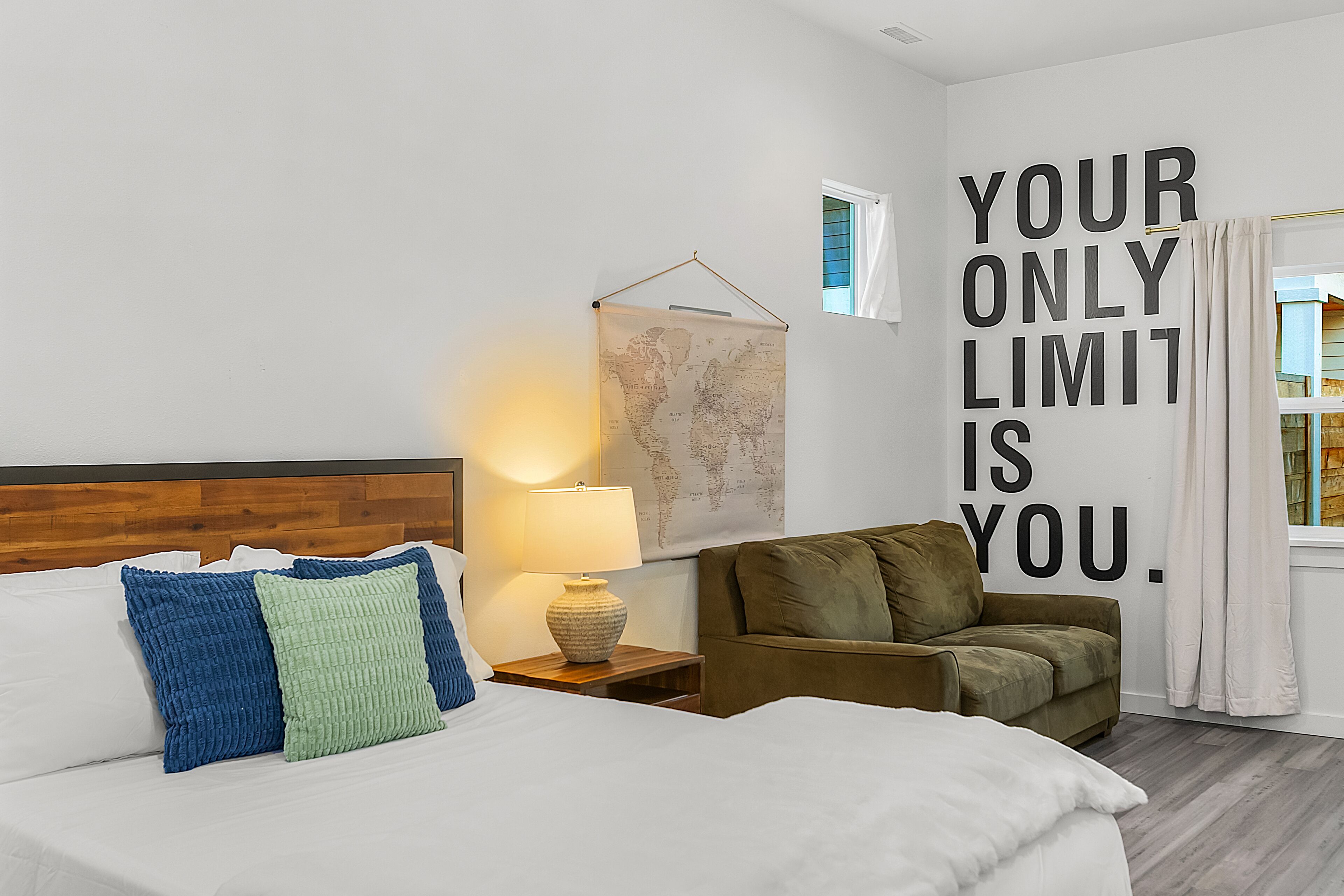 A hotel room with a bed, a nightstand with a lamp, a sofa, a world map on the wall, and a motivational quote.