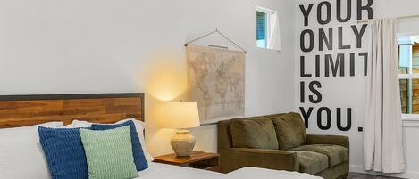 A hotel room with a bed, a nightstand with a lamp, a sofa, a world map on the wall, and a motivational quote.