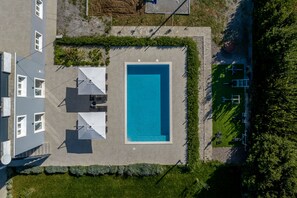 An aerial view of a modern building with a rectangular swimming pool, surrounded by a well-maintained lawn and a patio area with umbrellas.