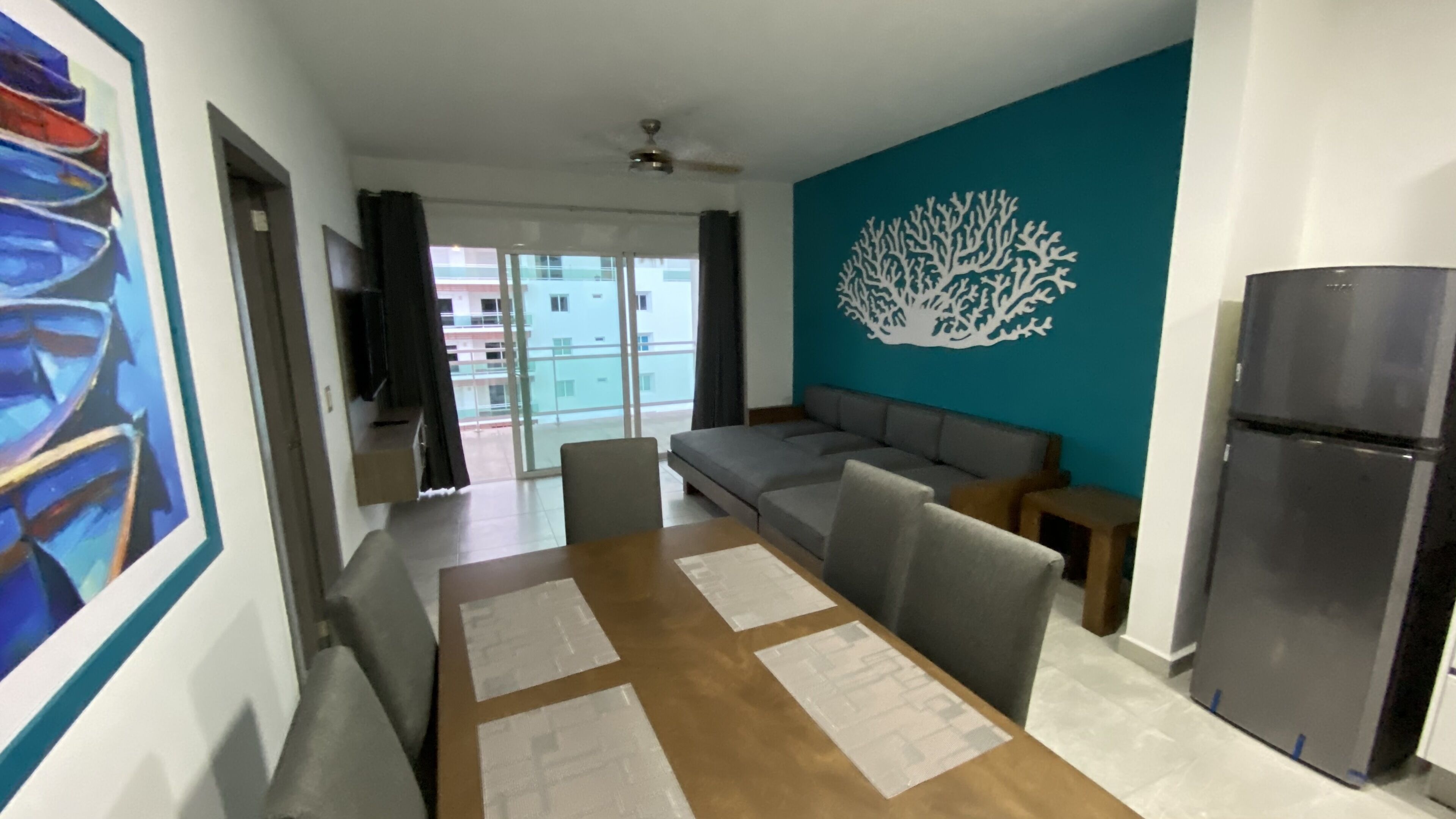 A modern living room with a sofa, a dining table with placemats, a refrigerator, and a wall with a coral reef mural.