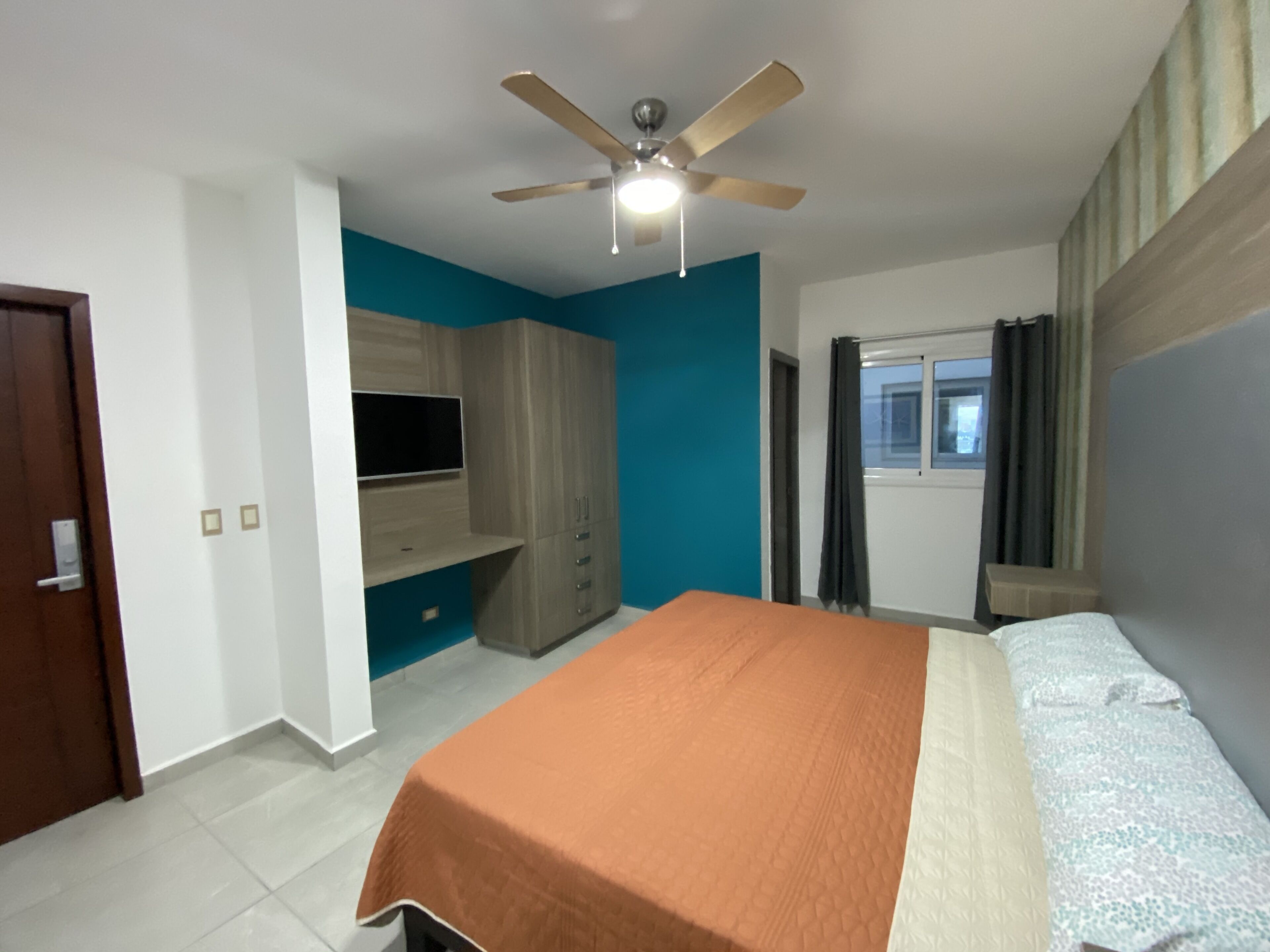 A bedroom with a bed, a ceiling fan, a TV mounted on a cabinet, and a window with curtains.