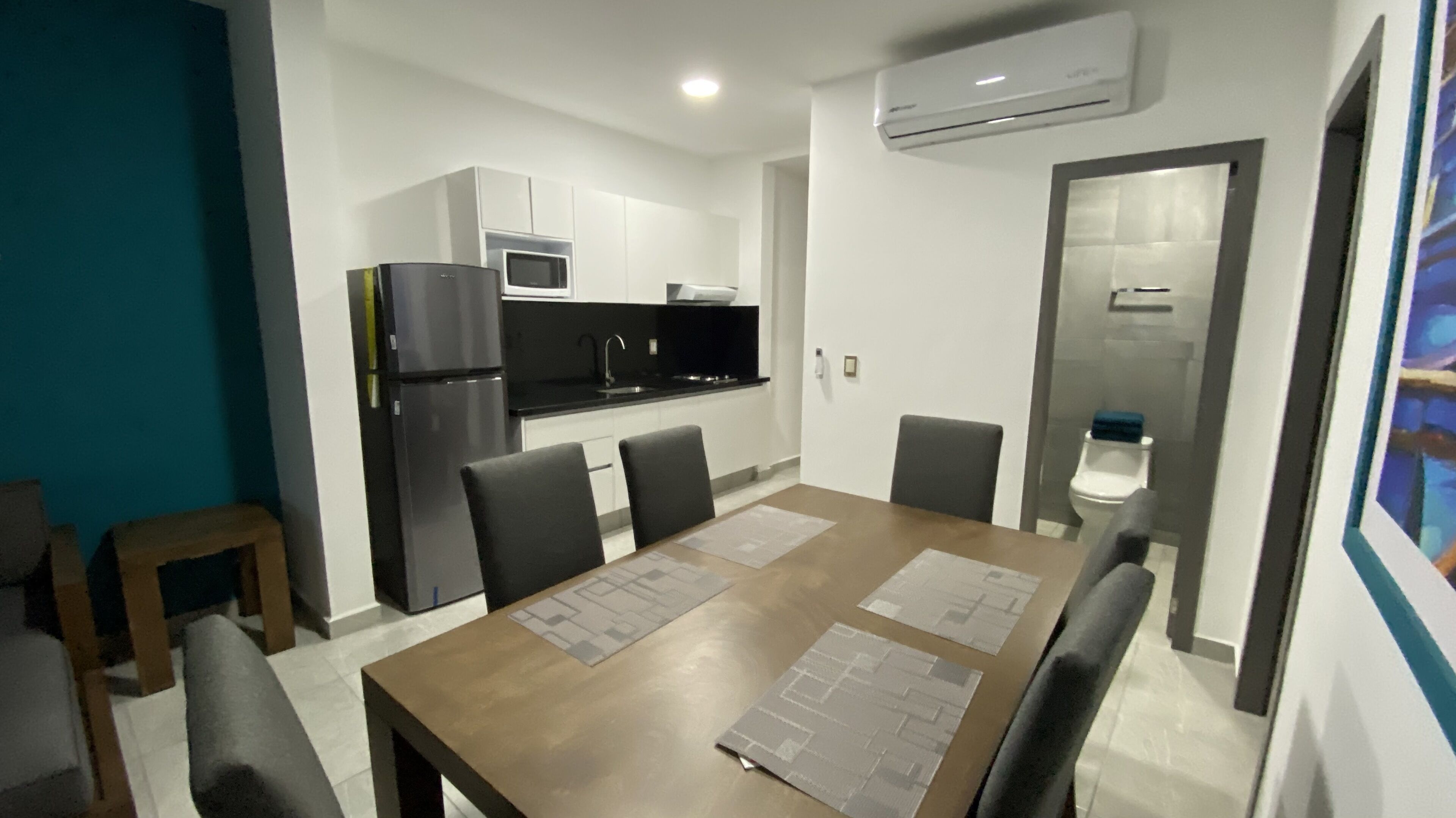 A modern kitchen with white cabinets, a black refrigerator, and a microwave. There is a dining table with four chairs and a small side table.