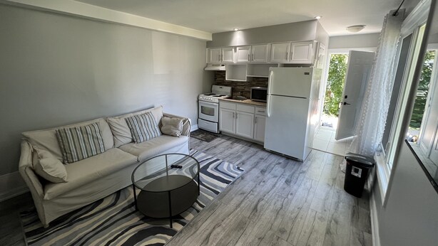 Cocina compacta con armarios blancos, un refrigerador y una estufa. Hay un sofá color beige con almohadones a rayas y una mesa de centro de vidrio.