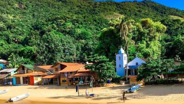 A beach with clear turquoise water, a few people swimming, and a small boat. There are buildings with colorful roofs in the background. - Pedaço de Céu Chalet: In the woods, 5 minutes from the beach (Paraty)