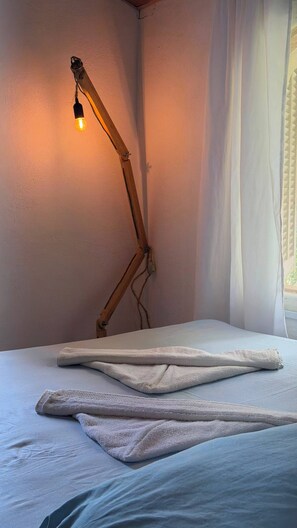 A bed with a white mattress and a folded white towel. A wooden floor lamp with a light bulb is positioned against the wall.