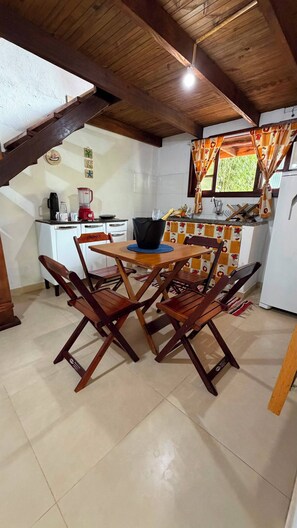 A dining area with wooden chairs and a table, a kitchen with a refrigerator, and a window with curtains.