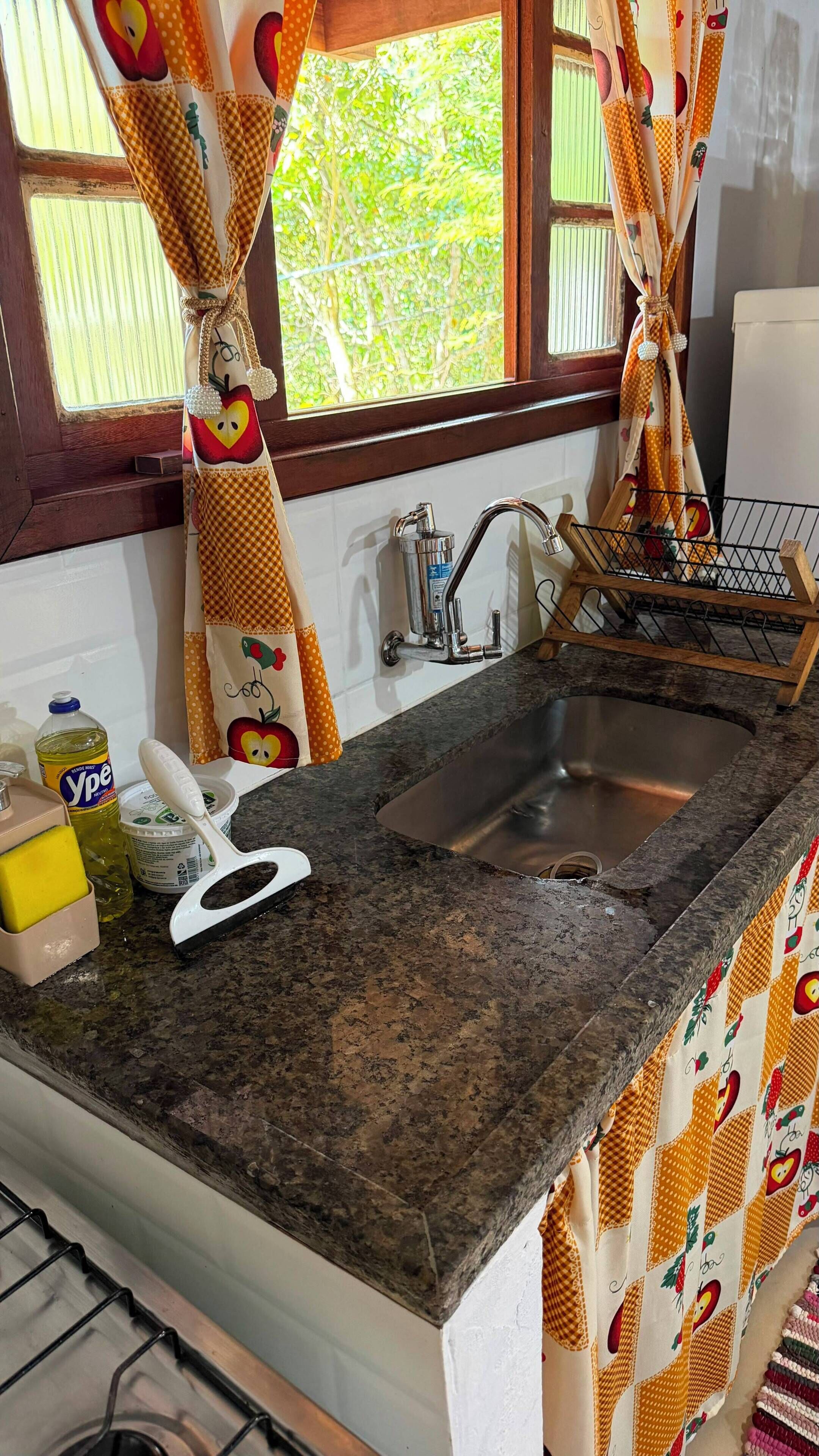 A kitchen with a granite countertop, stainless steel sink, and a window with curtains.