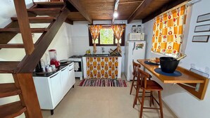 A compact kitchen with a gas stove, a microwave, and a refrigerator. There is a wooden dining table with two chairs and a black pot on it. - Pedaço de Céu Chalet: In the woods, 5 minutes from the beach (Paraty)