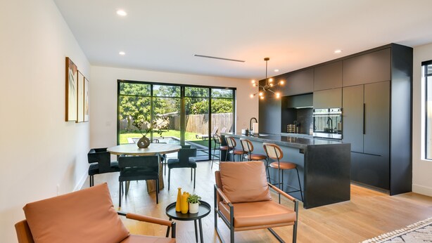 A modern kitchen with dark cabinetry, a central island, and a dining area with leather chairs and a round table.