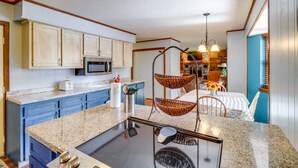Una cocina con encimeras de granito, armarios blancos, microondas y una silla colgante.
