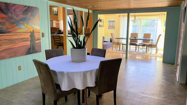 Uma sala de jantar com uma mesa redonda, cadeiras e um vaso de plantas. Há uma pintura na parede e vista da cozinha e da sala de jantar através de uma porta aberta.