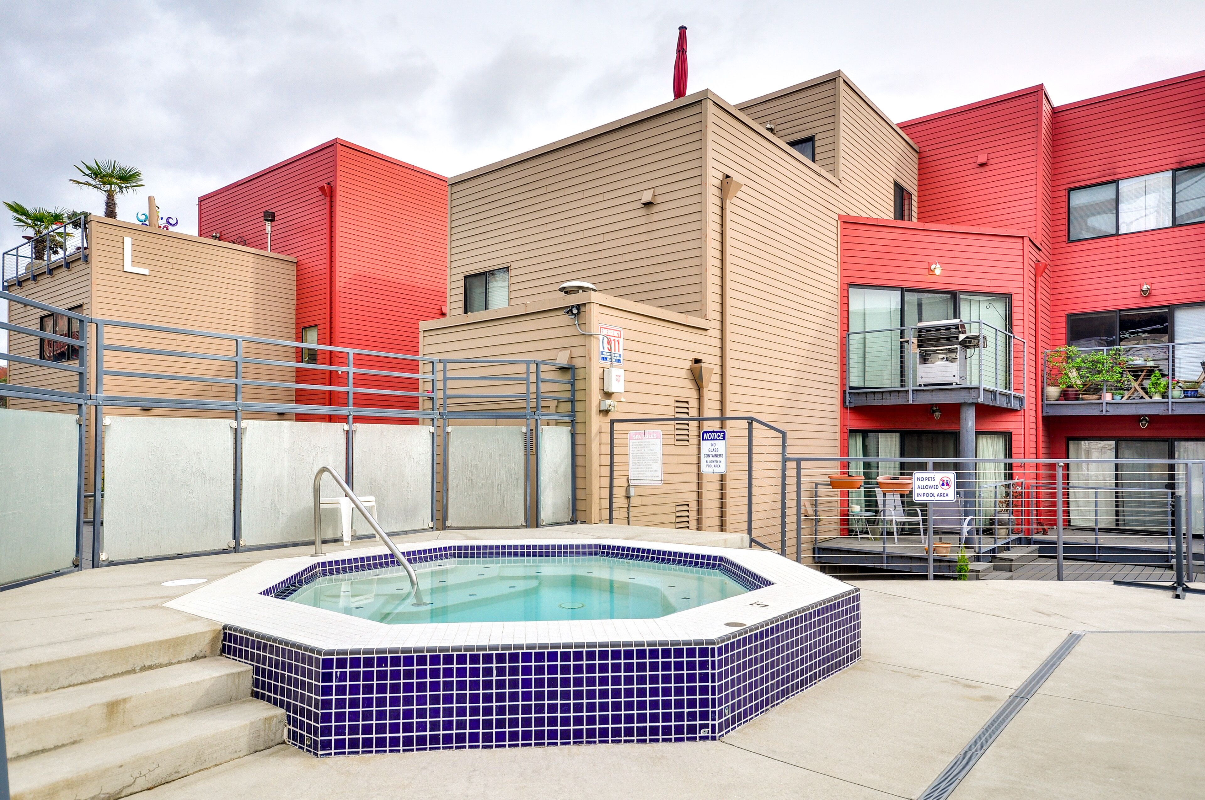 A swimming pool with a hot tub, surrounded by a building with red and beige facades, and a staircase leading to an upper level.