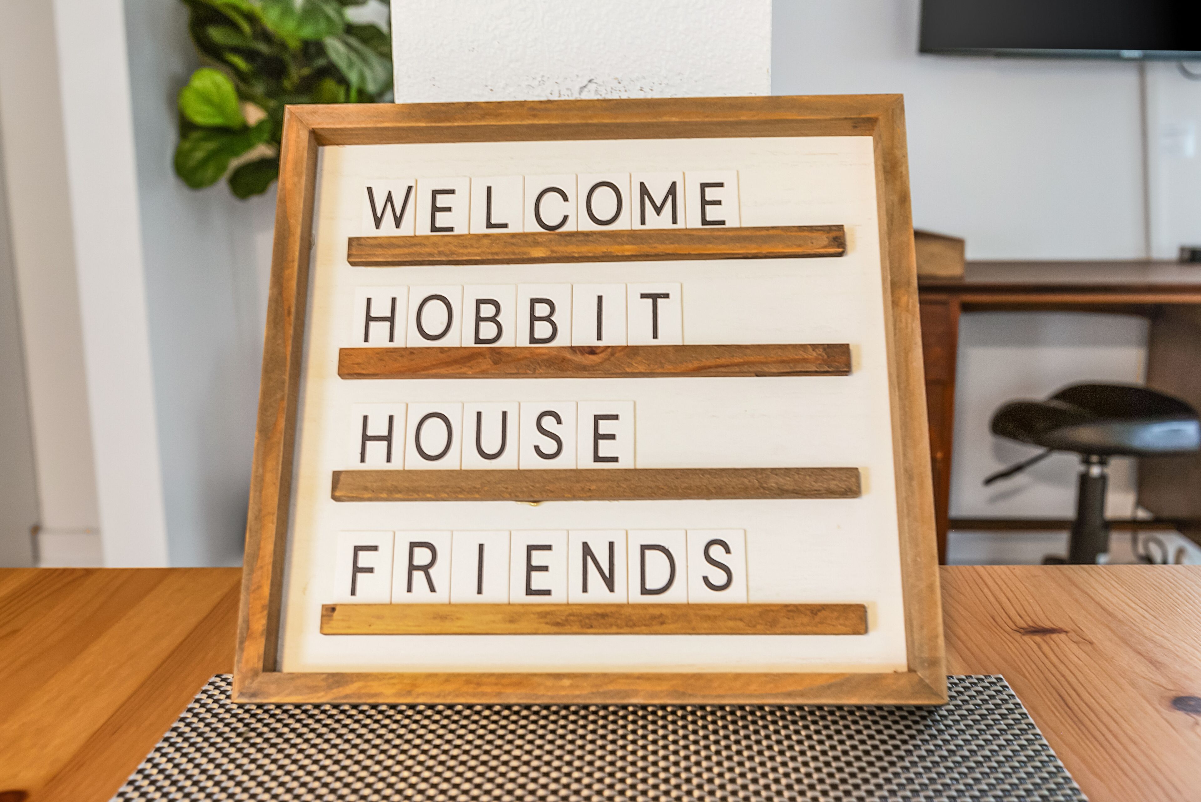 Un cartell de benvinguda emmarcat amb la frase "WELCOME HOBBIT HOUSE FRIENDS" damunt d'una taula de fusta.