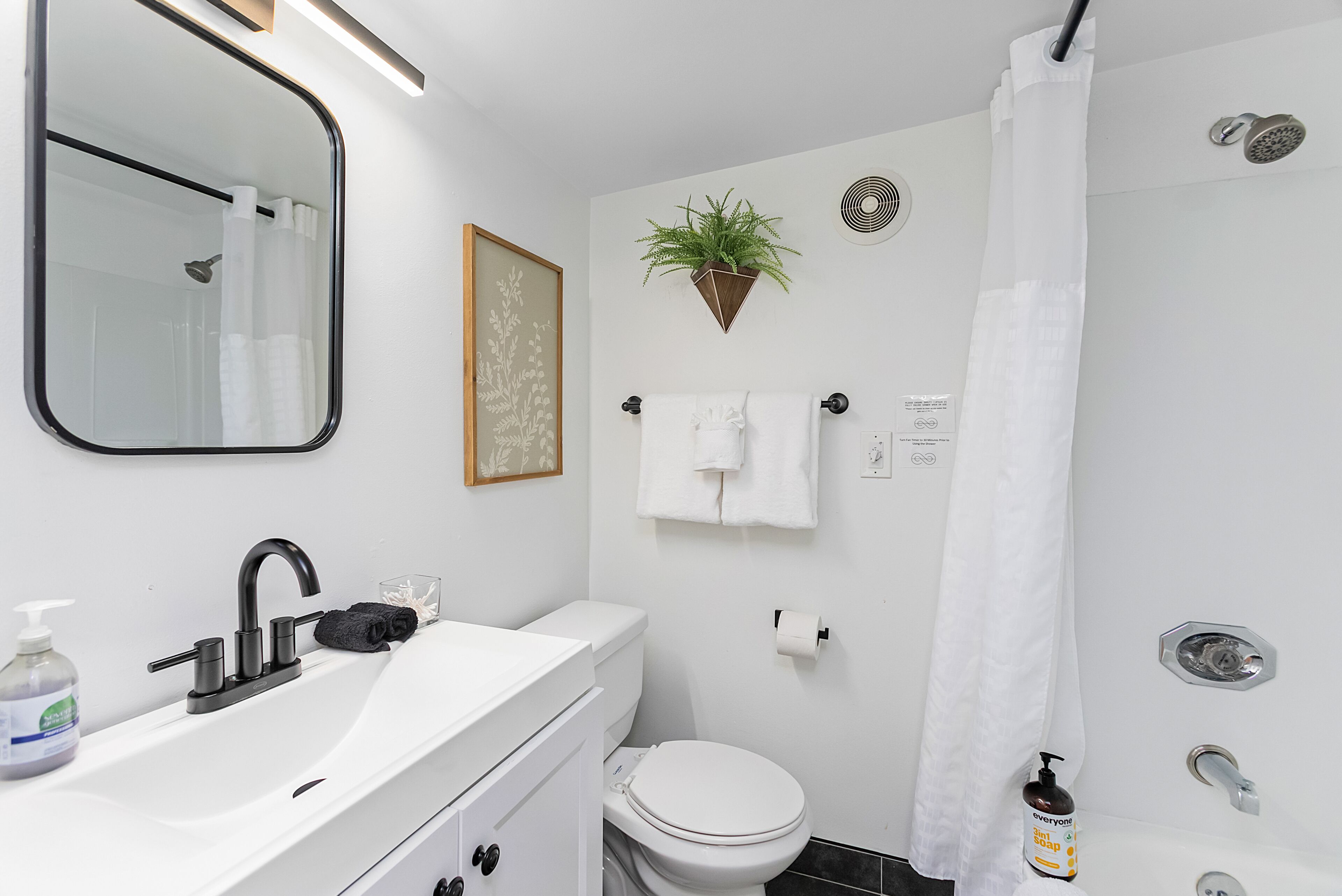 Un baño moderno con un lavamanos blanco, grifo negro y un espejo. Hay un inodoro, una ducha con cortina blanca y una planta en maceta en la pared.
