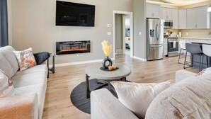 A modern living room with a fireplace, a flat-screen TV, and a kitchen in the background.