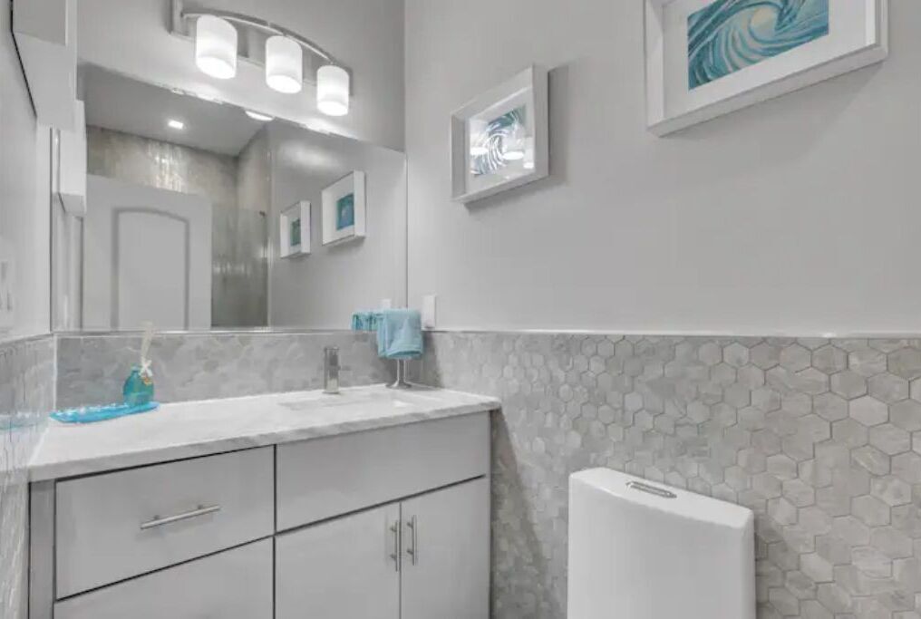 A bathroom with a white vanity, a mirror with lights, a hexagonal patterned wall, and a framed artwork.