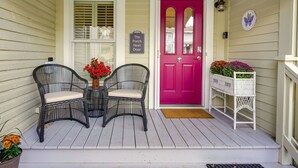Een veranda met twee rieten stoelen, een tafel met rode bloemen, een roze deur en een witte bank met bloemen.