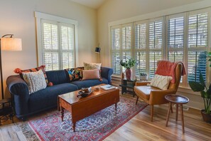 Un salon avec un canapé bleu foncé, une table basse en bois et un tapis à motifs. De grandes fenêtres sont équipées de stores, et on y trouve une lampe sur pied ainsi qu’une plante en pot.