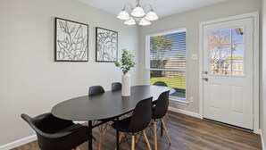 Uma área de refeições com uma mesa redonda, cadeiras pretas, um vaso branco com uma planta e duas obras de arte emolduradas na parede.