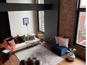 Una sala de estar moderna con un sofá, una mesa de centro y una alfombra. Se observa una pared de ladrillo y una ventana con vistas a edificios.