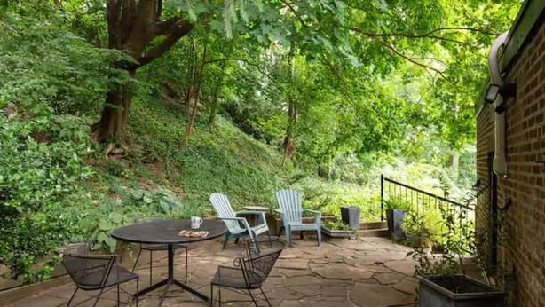 Une terrasse extérieure avec une table en pierre et des chaises en métal, entourée d’une végétation luxuriante et d’arbres.