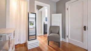 Un pasillo con piso de madera, una puerta blanca, una silla gris y un espejo que refleja una habitación con ventana.