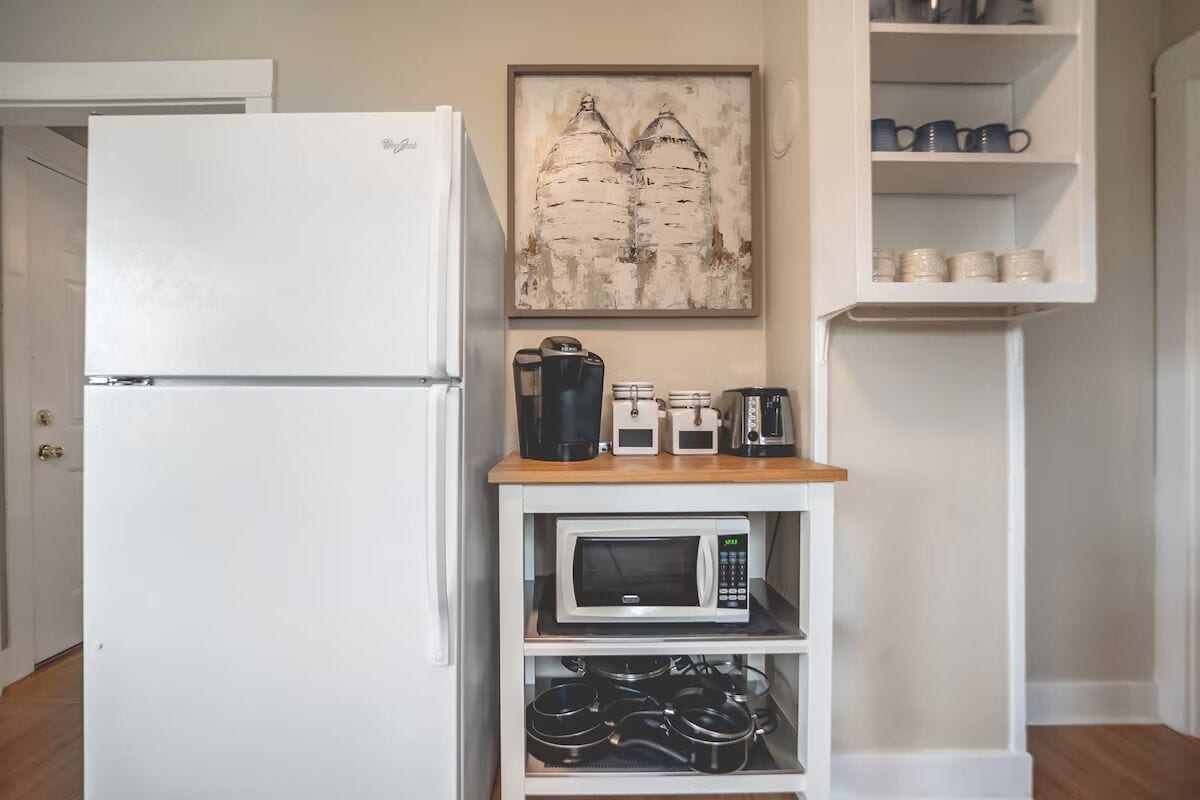 A kitchen with a white refrigerator, a microwave, a coffee maker, and a kettle on a wooden shelf.