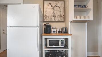 A kitchen with a white refrigerator, a microwave, a coffee maker, and a kettle on a wooden shelf.