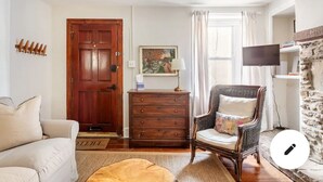 Una sala de estar con puerta de madera, sofá, sillón, cómoda, un cuadro, lámpara y televisor.