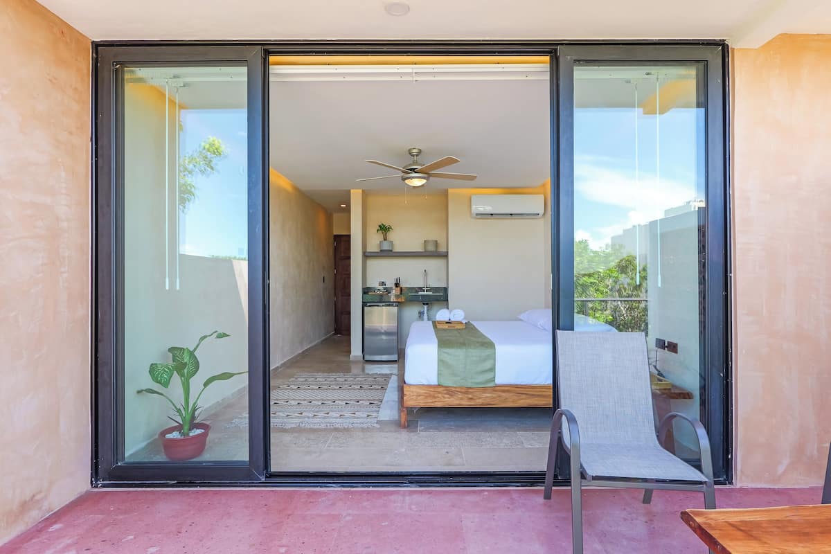 A bedroom with a bed, a chair, a table, a ceiling fan, and a small kitchenette.